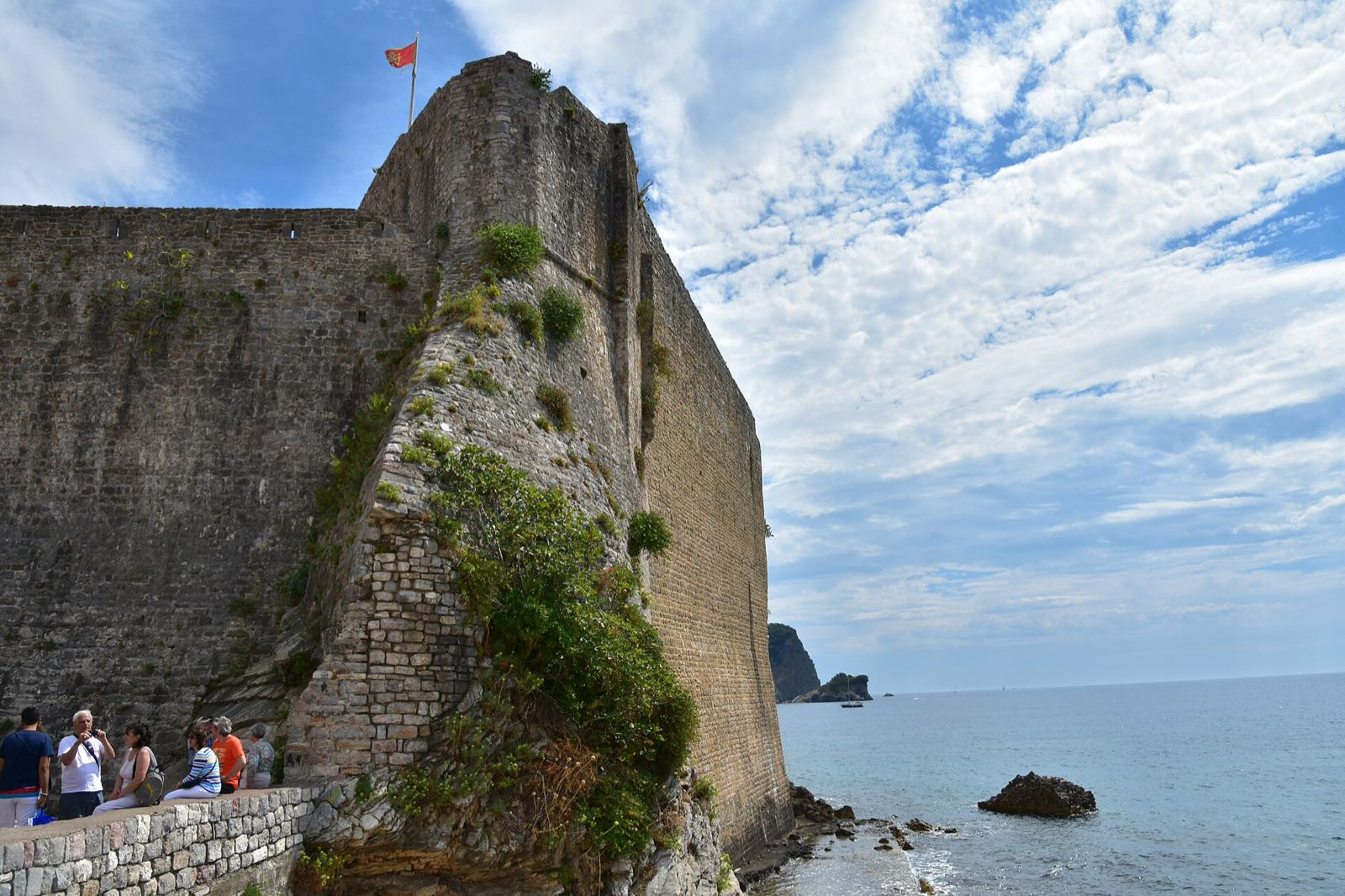 The south face of Budva's sea walls dropping into the Adriatic, the Montenegrin flag flying from the bastion